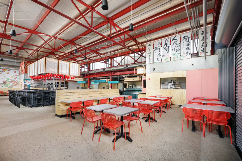 Interior of an industrial-style restaurant with red metal ceiling trusses, wood-topped tables, red chairs, and hanging banners with Chinese calligraphy.