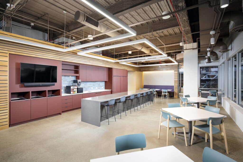 Office break room with a long kitchen island, bar stools, square dining tables, and a wall-mounted television.