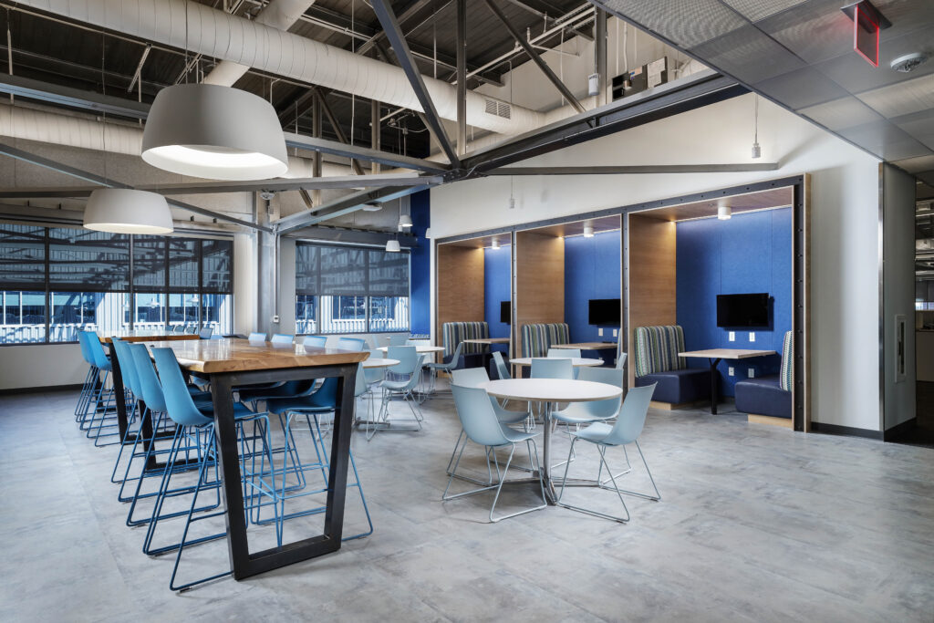 Modern office break room with a high communal table and blue bar stools, plus several small round cafe tables and individual booth seating with blue walls.