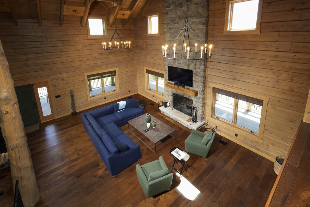 High-angle view of a living room with log walls, a stone fireplace, a blue sectional sofa, and two green armchairs.