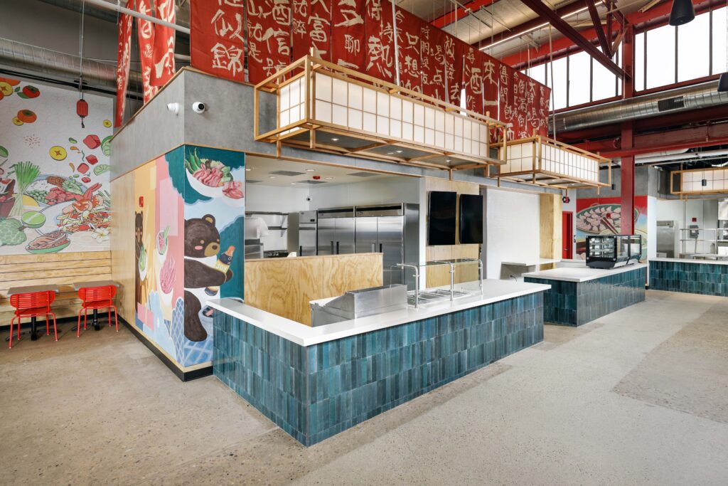 An open-concept restaurant service counter with teal tile, light wood accents, and red banners with Asian characters hanging above.