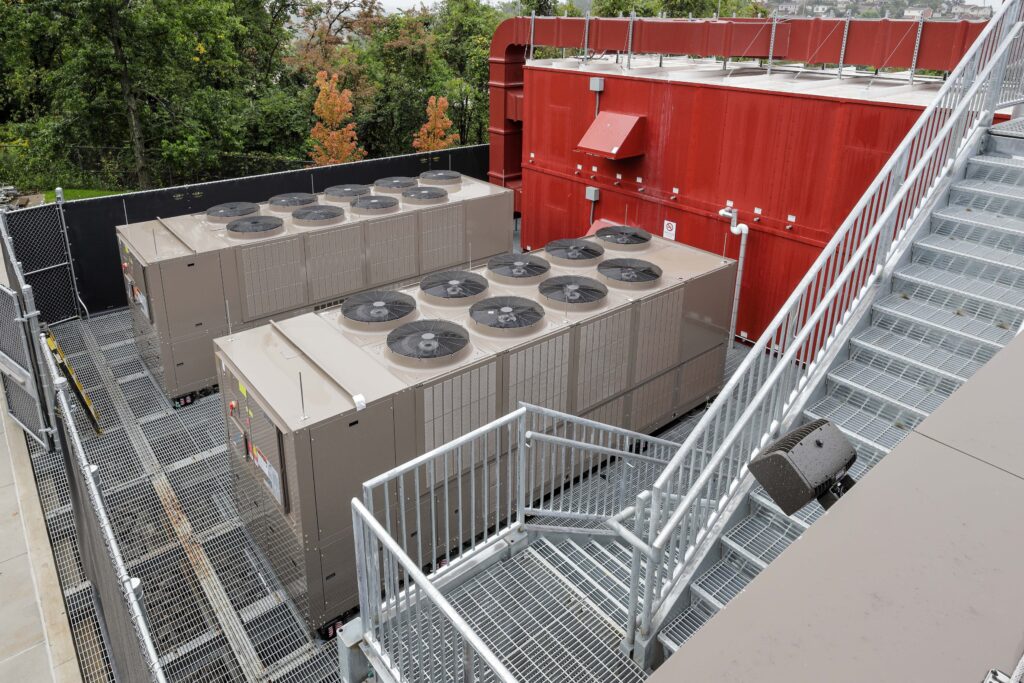 Two large industrial HVAC units on a grated rooftop platform next to a red mechanical housing and metal stairs.