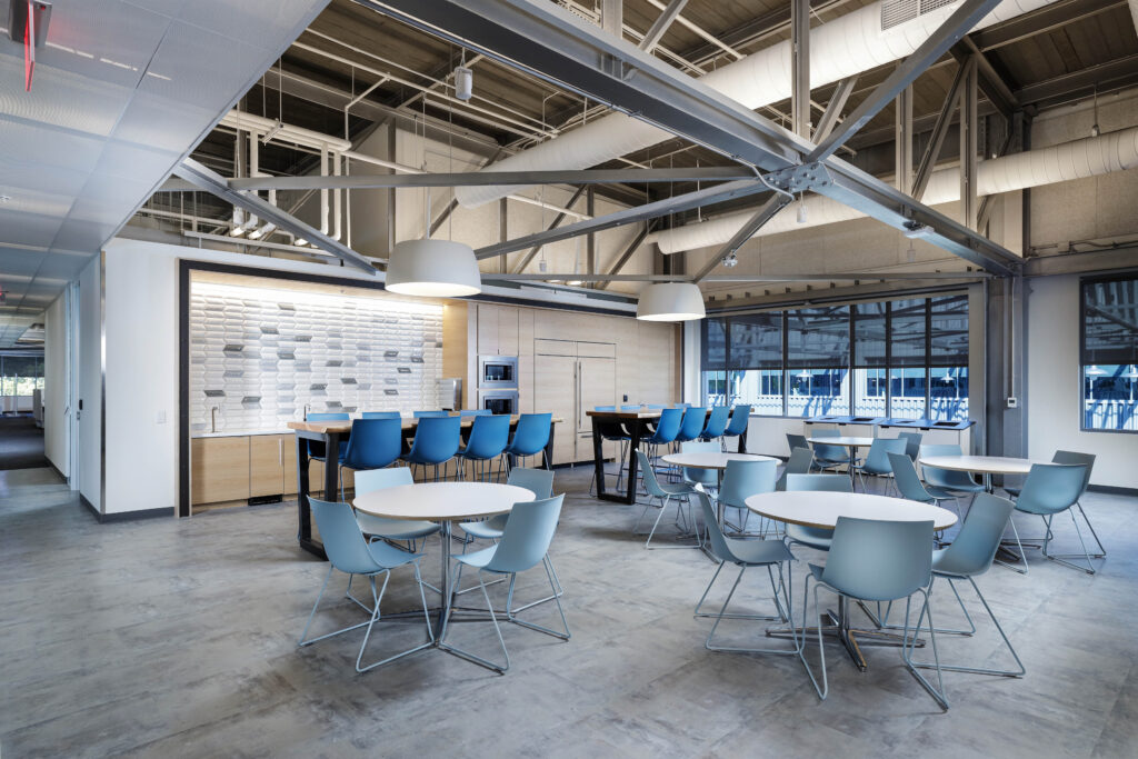Modern office breakroom with blue chairs, round tables, a long counter with stools, and an exposed ceiling structure.
