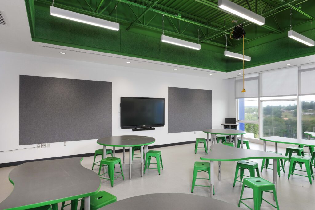 A classroom with grey tables, green stools, a large wall-mounted monitor, and a green industrial ceiling.