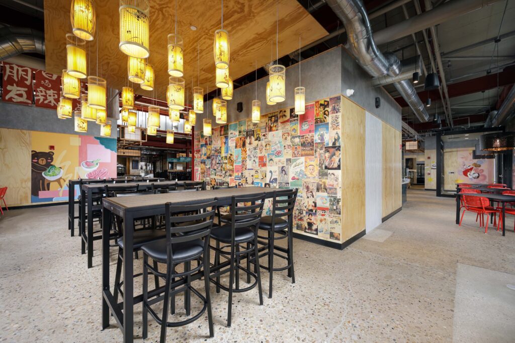 A dining area with high-top tables, black bar stools, and cylindrical pendant lights, featuring a wall covered in various posters.