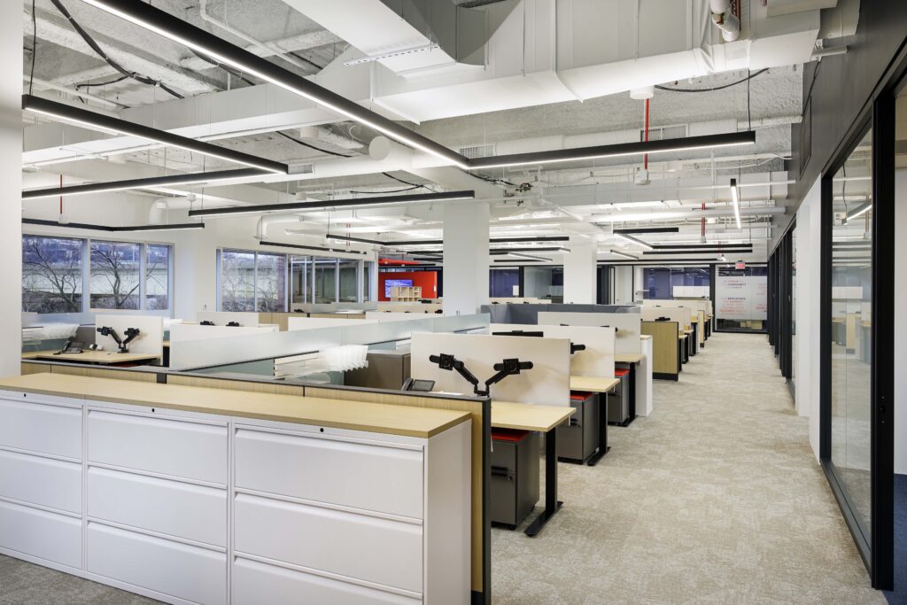 An open-plan office space with rows of cubicle desks and white storage cabinets.