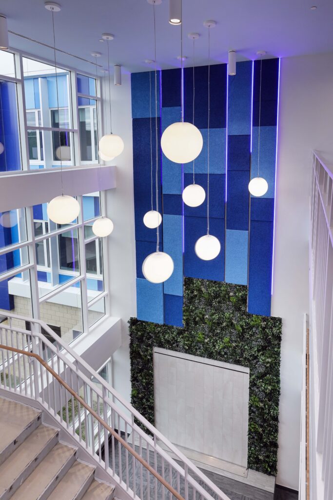 A high-ceilinged atrium with a staircase, spherical pendant lights, blue acoustic wall panels, and a vertical garden.