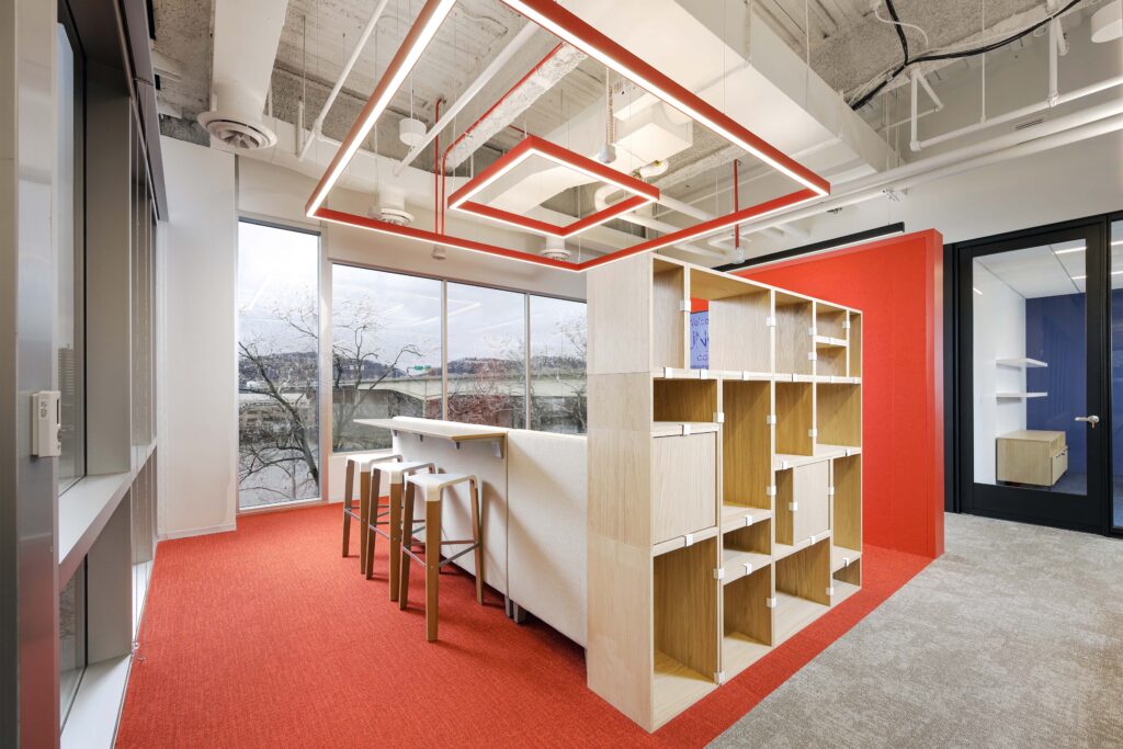 Modern office space featuring a tall wooden shelving unit, a high-top table with three stools, and a bright red carpet, with a view of a bridge through a large window.