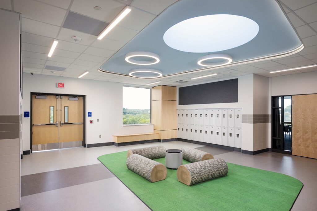 A school hallway with a seating area featuring four log-shaped benches on a green rug, white lockers, and a double door exit.