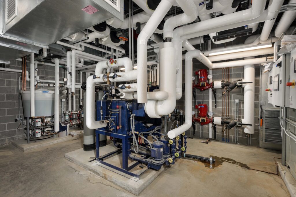 Mechanical room with insulated white pipes, a blue industrial chiller unit, and red water pumps against a concrete block wall.