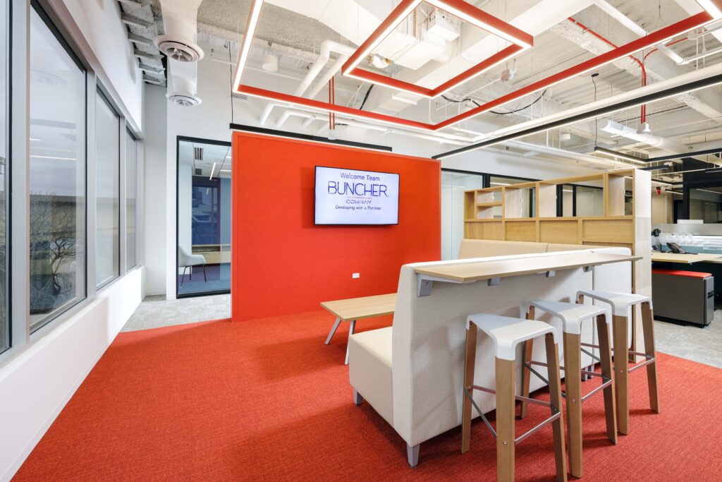 A modern office lounge with a bright red wall, featuring a TV screen displaying “Welcome Team BUNCHER COMPANY,” a high-top table with stools, and orange carpeting.