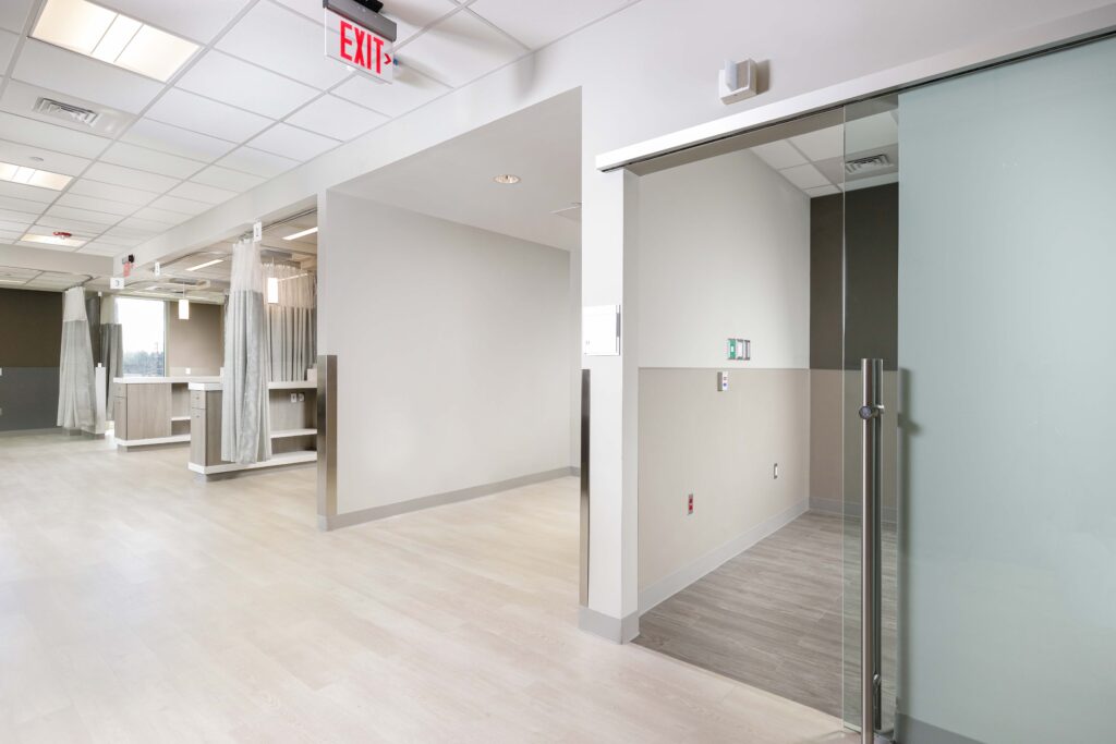 Hospital hallway with a patient exam room featuring a privacy curtain and a glass-paneled entry door.