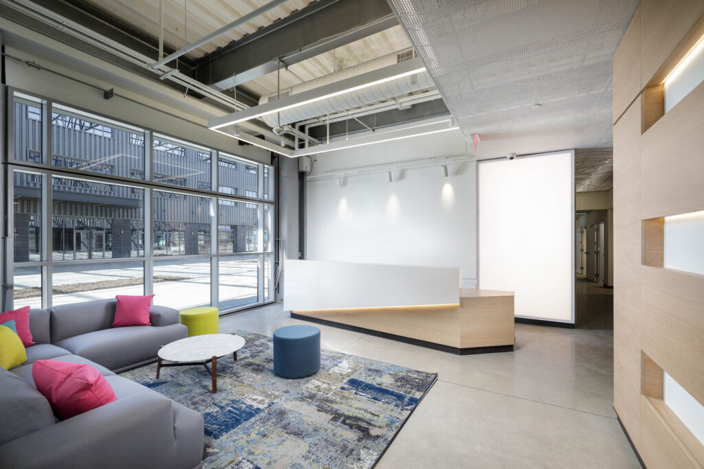 Modern office lobby with a reception desk, a gray sofa with colorful accent pillows, a coffee table, and large windows.