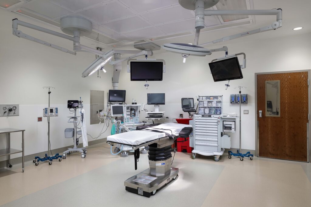 An empty operating room with a surgical table in the center, surrounded by medical equipment, monitors, and overhead surgical lights.