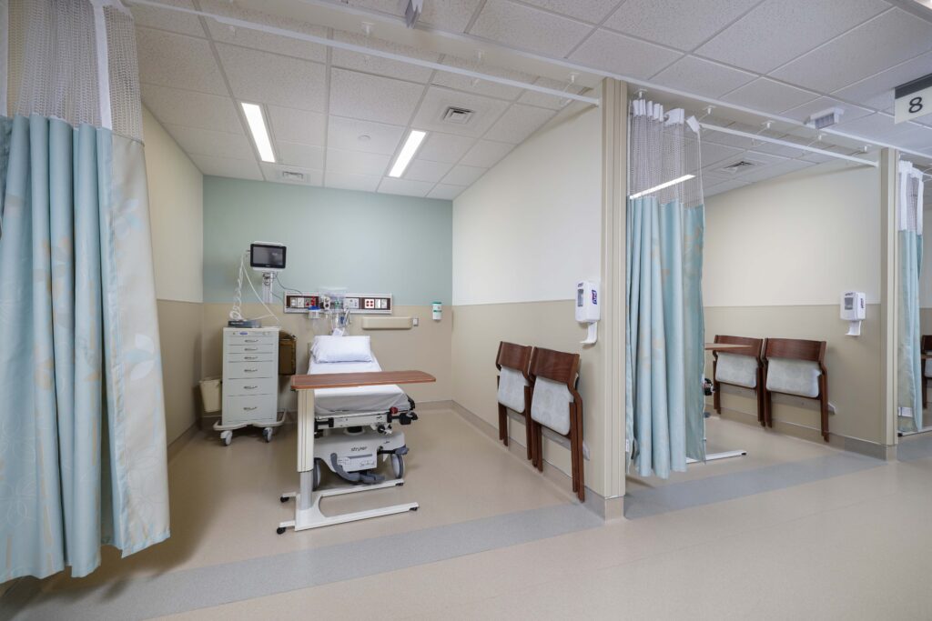 A hospital patient room with an adjustable bed, rolling medical cart, and two chairs.