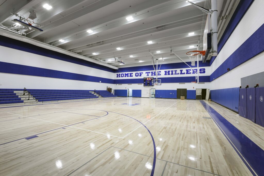 Indoor basketball court with polished wood floors, blue bleachers, and the text “HOME OF THE HILLERS” on the far wall.