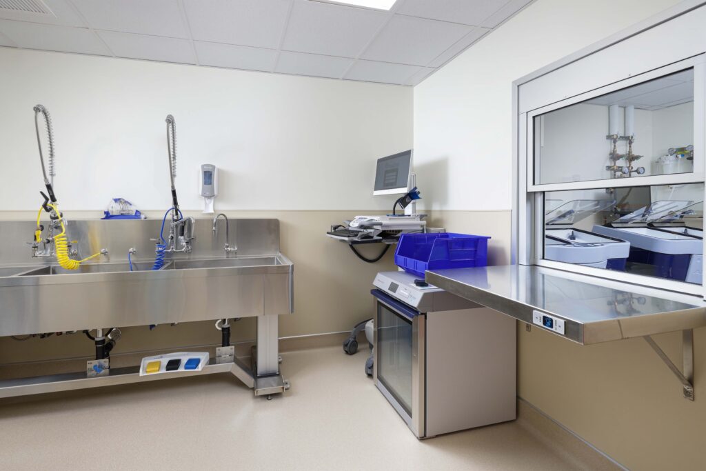 A medical or veterinary exam room featuring a stainless steel scrub sink, a computer workstation, and an under-counter medical refrigerator with a blue bin on top.