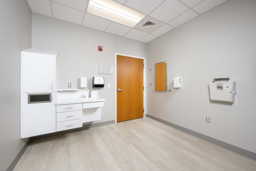 A medical exam room featuring a sink, cabinets, a closed wooden door, and a wall-mounted infant changing station.