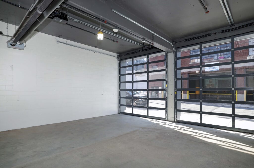 An empty garage interior with a concrete floor, white block wall, and two large, glass-paneled rolling doors opening to a street.