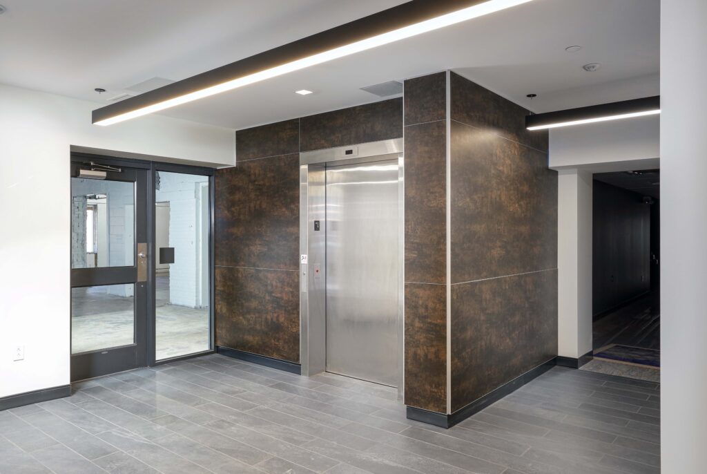Stainless steel elevator in a lobby with dark, textured wall panels and gray tile flooring.