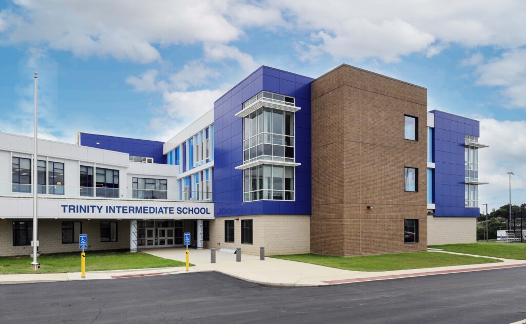 Exterior of Trinity Intermediate School, a multi-story building featuring blue panels, tan brick, and a glass-enclosed entrance area.