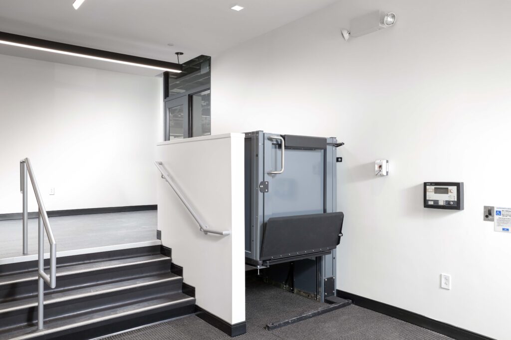 A vertical wheelchair lift installed next to a short flight of stairs in a modern building hallway.