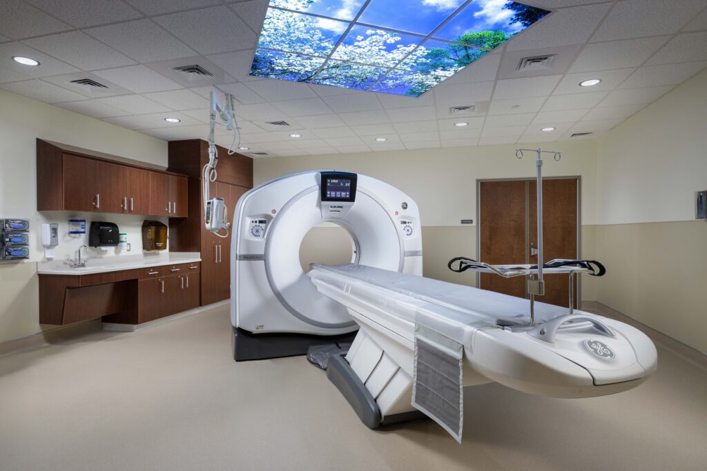 A CT scanner in a medical imaging room with a patient bed extended in front of the gantry and a nature-themed light panel on the ceiling.