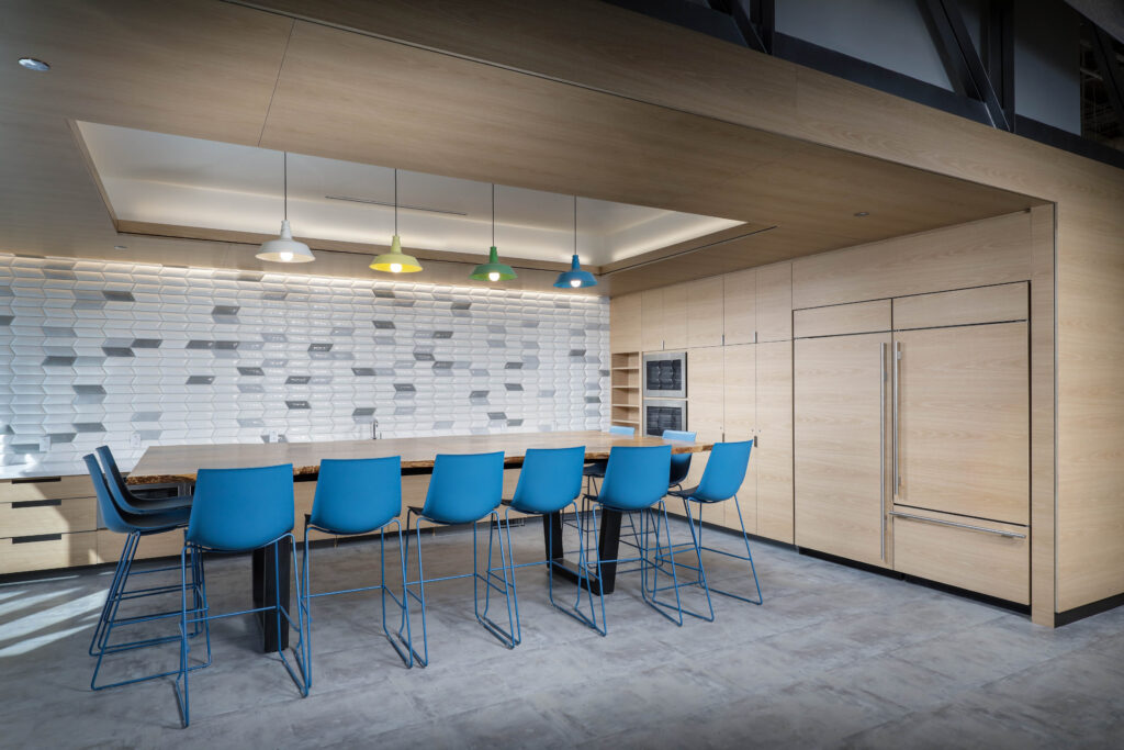 A wooden kitchen island with six blue bar stools, illuminated by four colorful pendant lights in front of a patterned white tiled wall.