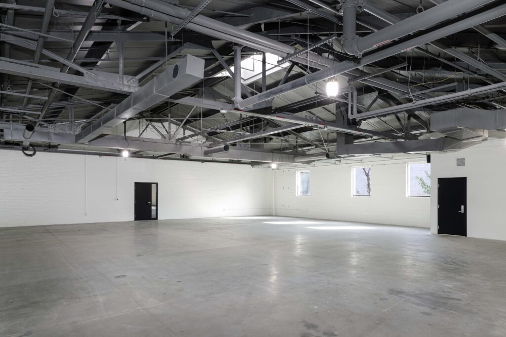 An empty industrial warehouse space with white brick walls, a concrete floor, and exposed metal rafters and piping on the ceiling.