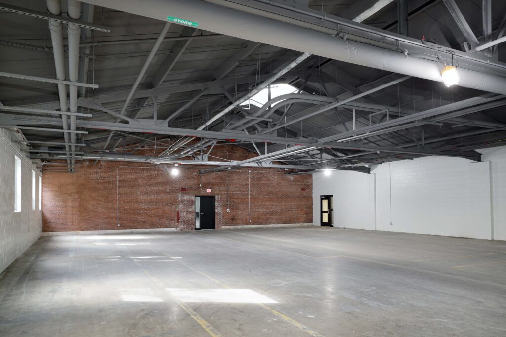 An empty industrial warehouse space with brick and white walls, exposed rafters, and concrete floors.