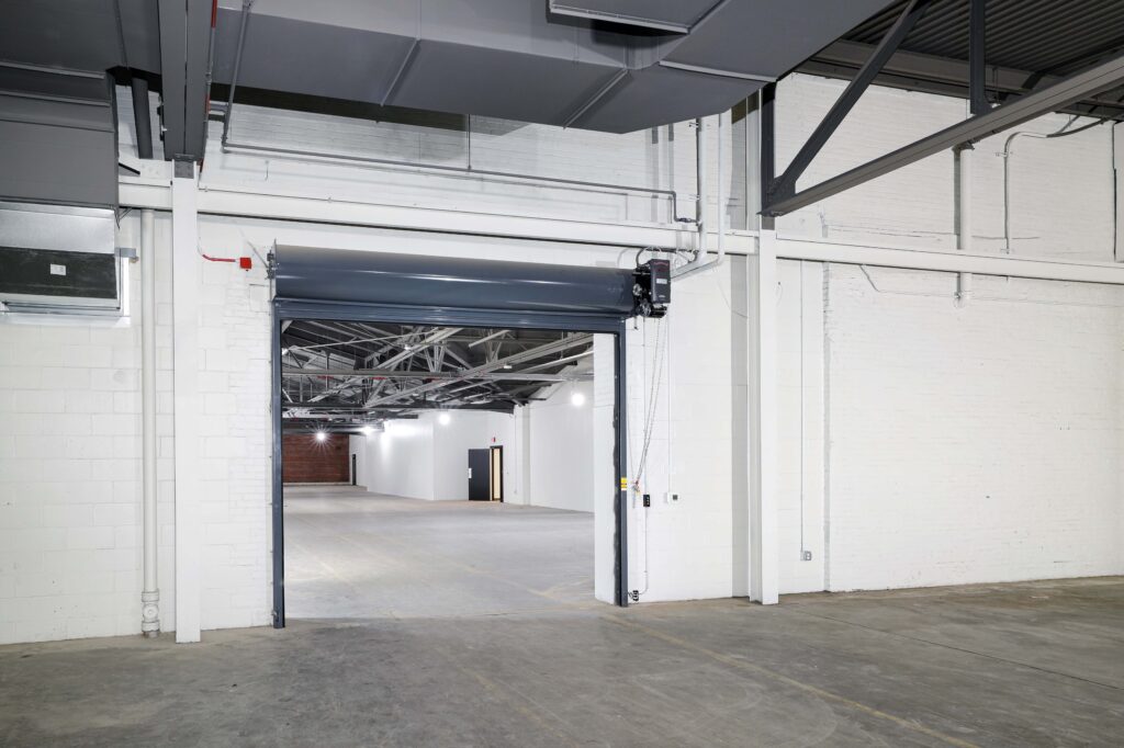 An open, gray roll-up industrial door leads from a foreground room into a larger, empty warehouse space with white brick walls.