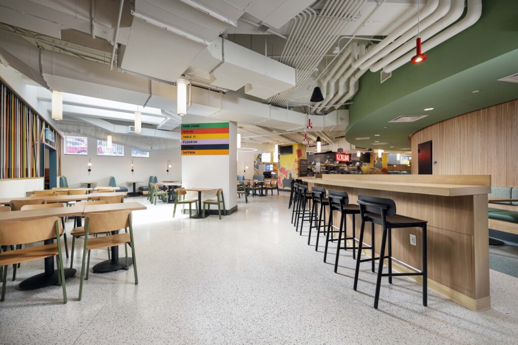 Modern food hall interior with a wooden bar counter, black stools, and surrounding tables and chairs. A central column displays a colorful list of restaurant names.