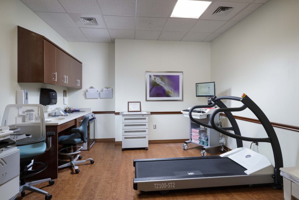 A medical examination room with a treadmill, a mobile cardiac monitoring station, a desk with office chairs, and a small medical cart.