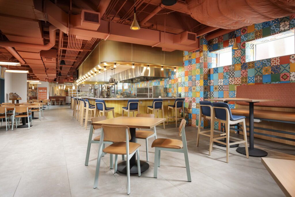 Modern dining area with a food service counter, colorful patterned wall tiles, and wood and metal tables and chairs.