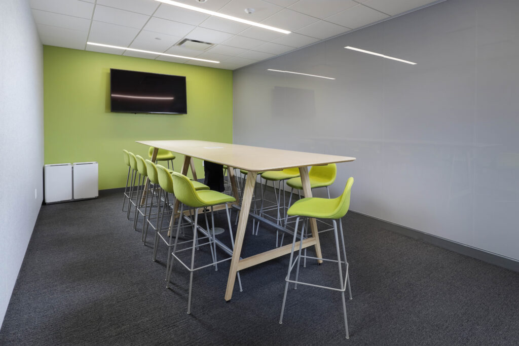 A tall wooden table surrounded by lime green bar stools in a meeting room with a wall-mounted flat-screen television.