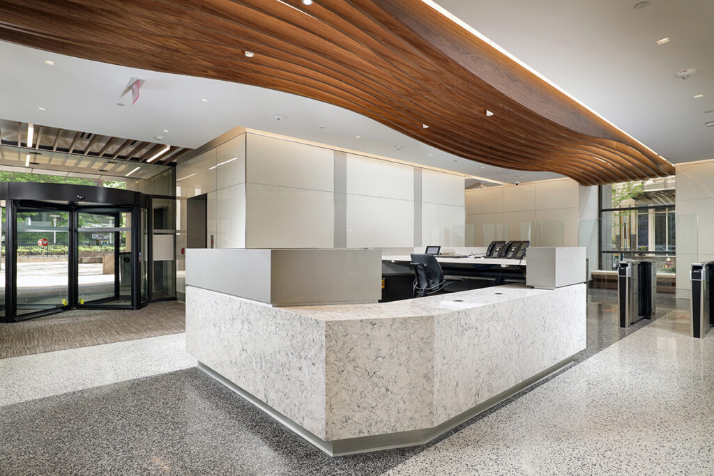 A modern building lobby featuring a large white marble-patterned reception desk and a prominent wooden ceiling structure with wavy, slats.