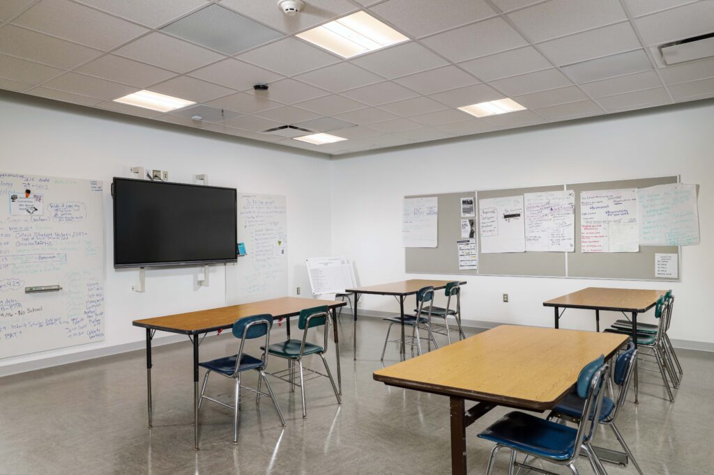 A classroom with three tables and blue chairs, a large digital display mounted on the wall, and multiple whiteboards covered in handwritten notes.