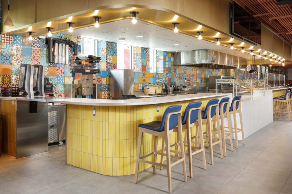 A food service counter with a yellow tiled base, a white top, and a row of blue stools, set against a wall of colorful patterned tiles.