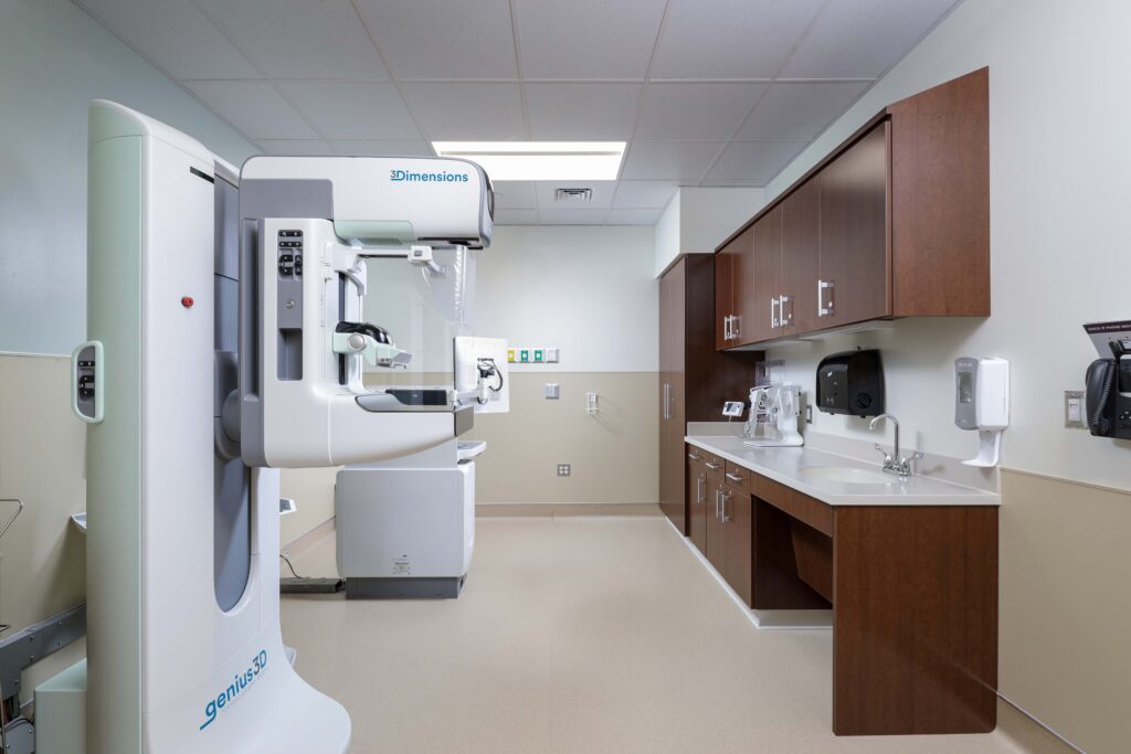A 3D mammography machine in a medical exam room with brown cabinetry and a sink.