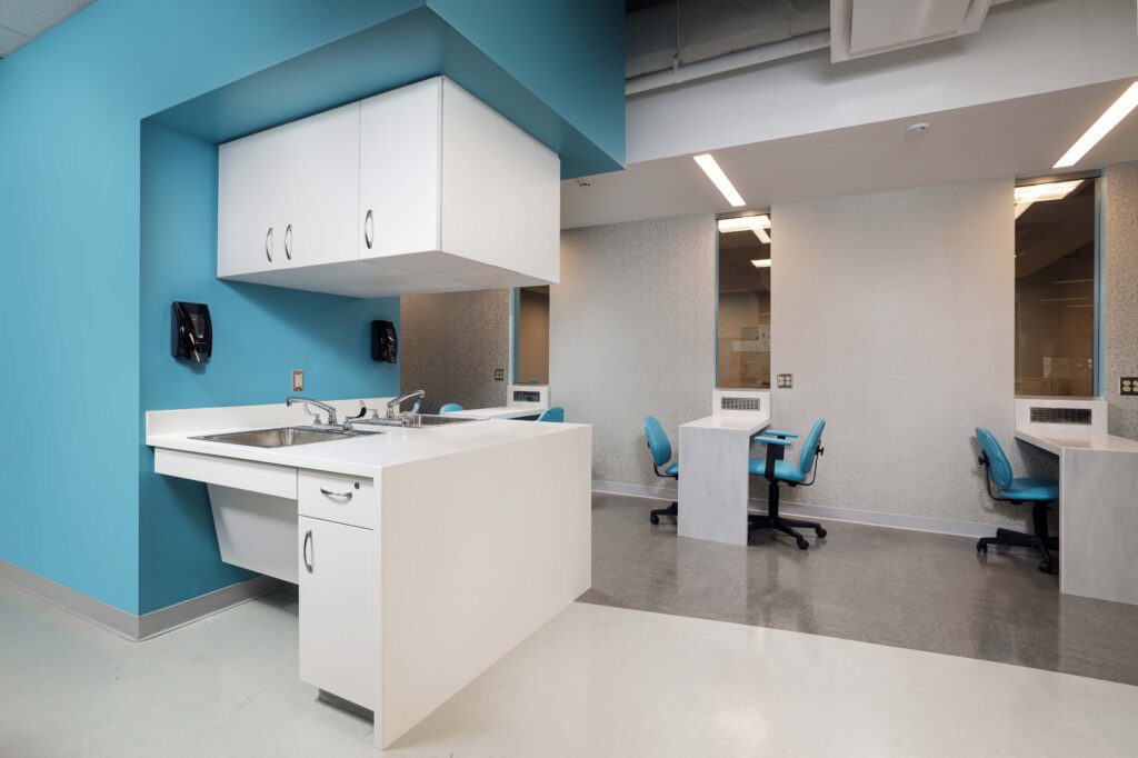 A clinical workspace with a white sink station, white cabinetry, and two desks with blue office chairs against light-colored walls.