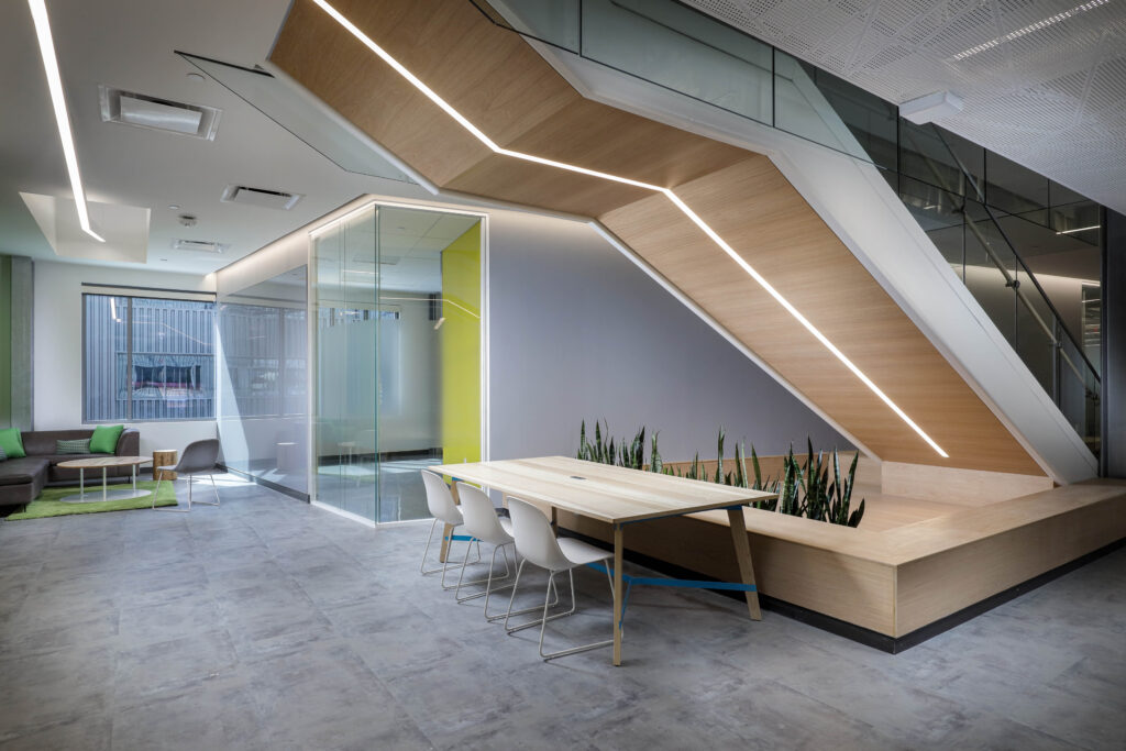 Office interior with a wooden staircase featuring integrated lighting, a meeting table with white chairs, and a glass-enclosed room with a seating area.
