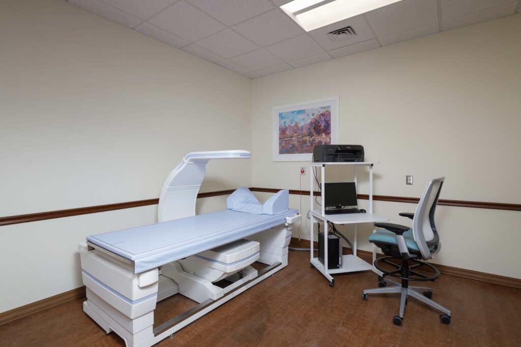 A medical examination room with a DXA bone density scanner, a computer workstation with a printer, and an office chair.