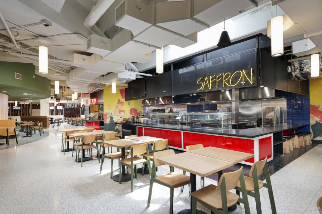 A cafeteria dining area with light wood tables and chairs, featuring a food service counter with a glowing yellow “SAFFRON” sign.