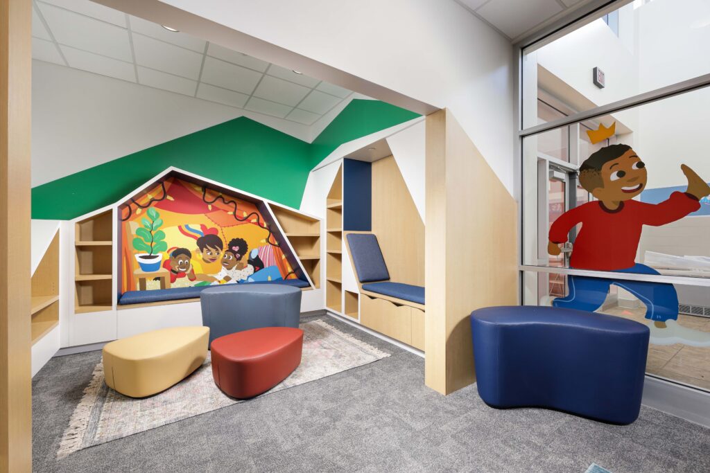 A children’s reading nook with colorful bench seating, abstract stools, built-in shelving, and a wall mural featuring children and a cartoon character on the glass.