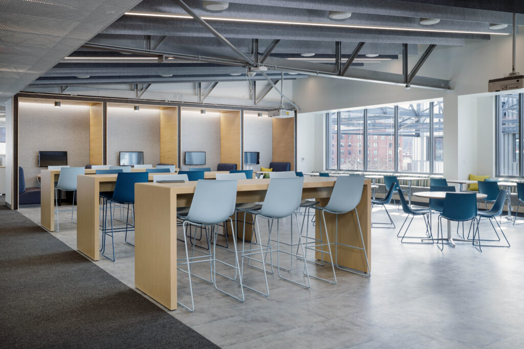 Modern office space with tall wooden desks, light blue stools, and individual computer workstations along the back wall.