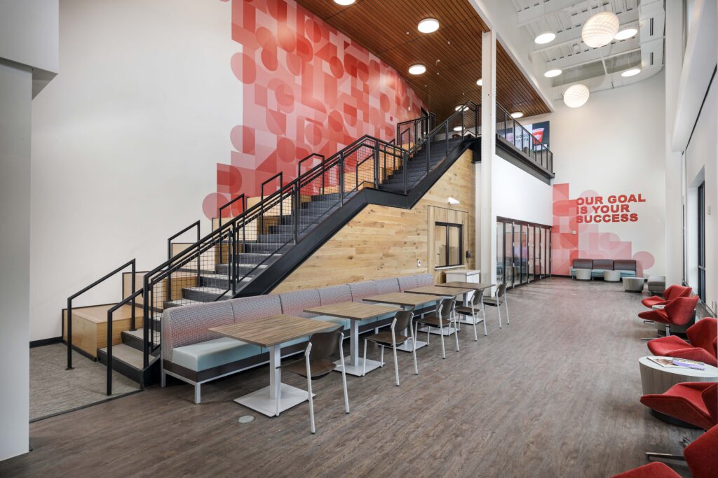 Modern office lobby with a staircase, rows of tables and chairs, and red geometric wall murals.