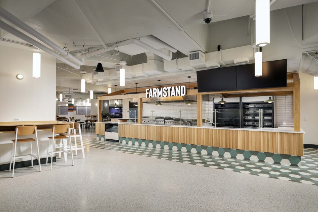 A modern cafe counter labeled “FARMSTAND” with a wooden front, glass food display, and high-top seating area.