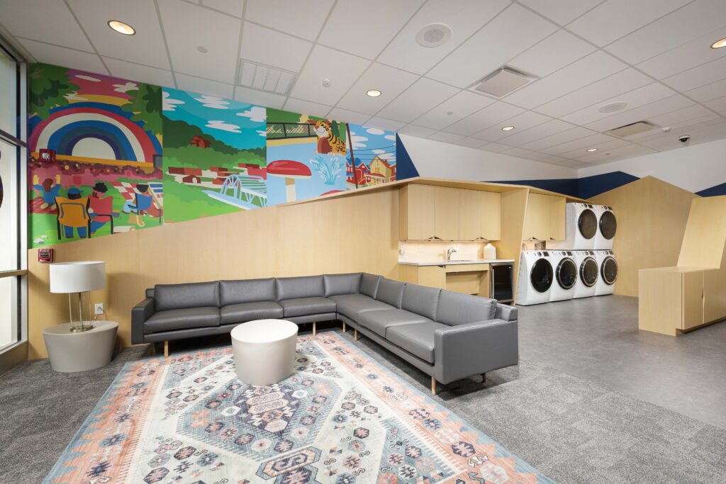 A lounge area with a gray sectional sofa, round coffee table, patterned rug, and a wall mural featuring a colorful park scene, with a laundry area containing two stacked sets of washers and dryers.