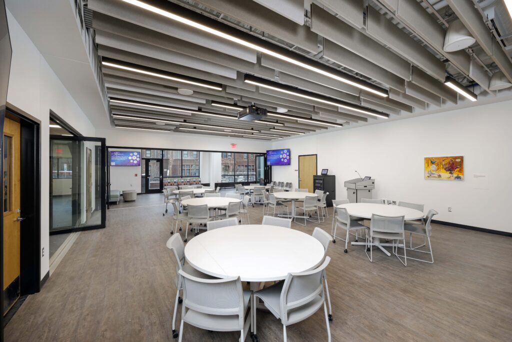 A large conference room with round tables, chairs, two wall-mounted televisions, and a podium.