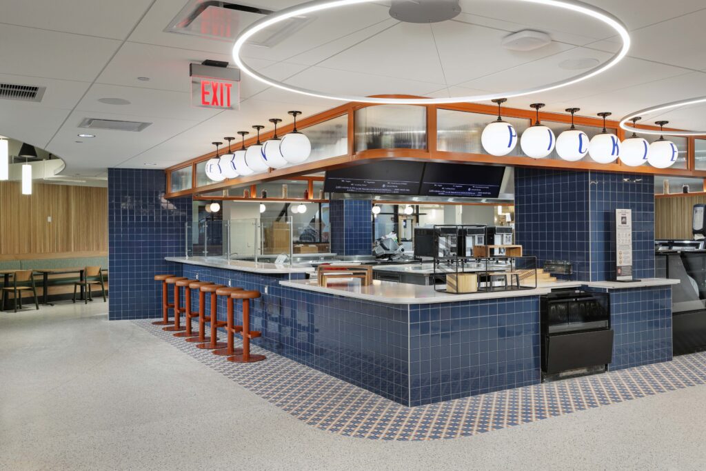 A modern cafeteria counter with blue tiled walls, high stools, and a ring-shaped pendant light.
